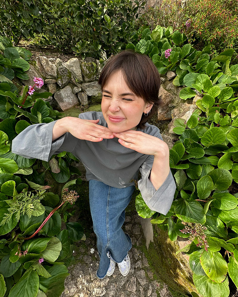 A full-body picture of Lera smiling, with flowers in the background. She has shoulder-length hair and is wearing a long-sleeve t-shirt and a denim skirt.